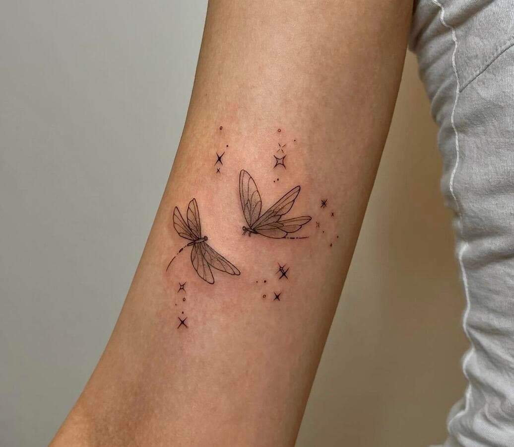 Two fineline dragonflies tattooed on an upper arm, surrounded by small scattered sparkles, all drawn in a single thin black line weight.